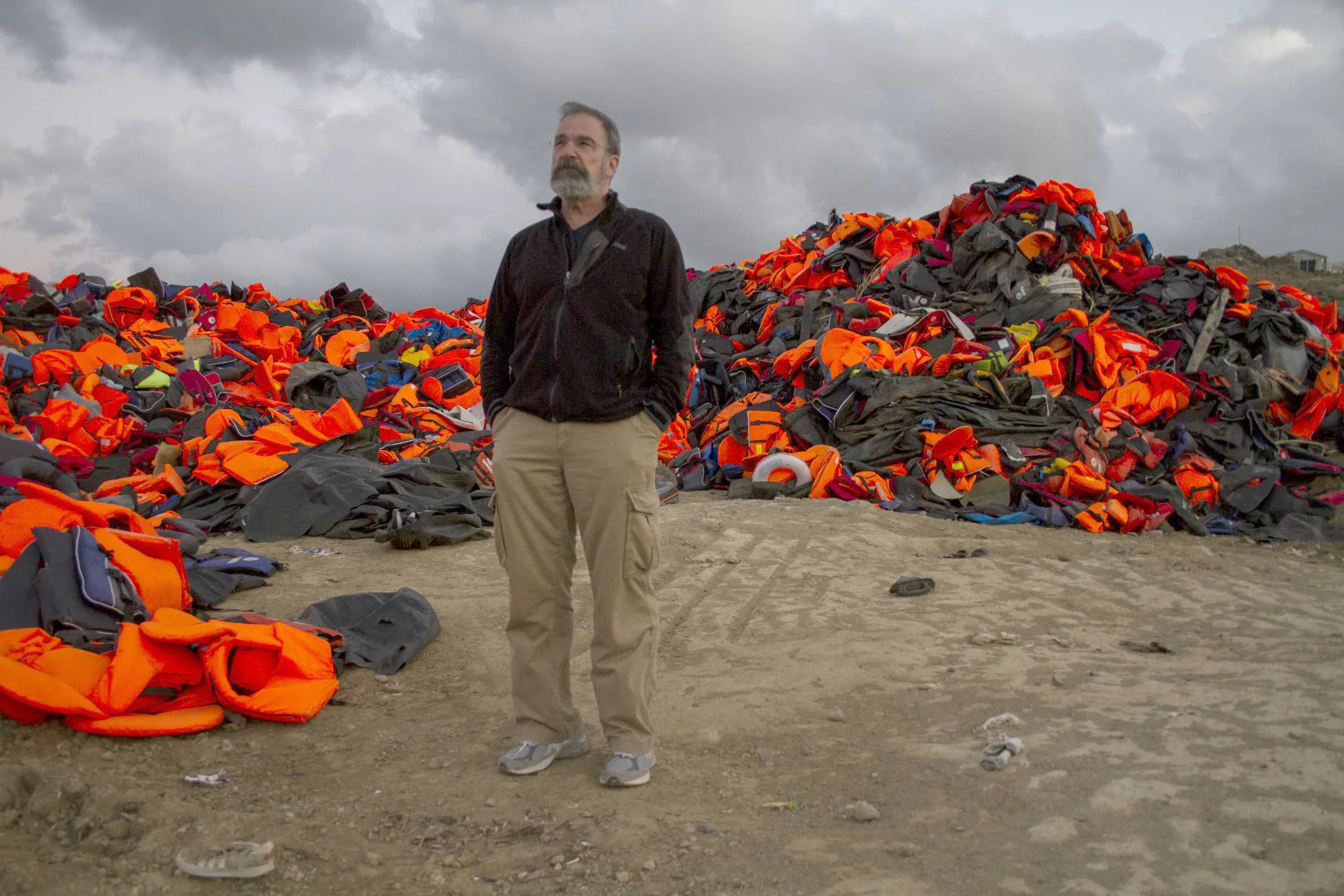Mandy Patinkin standing before a massive pile of colorful life jackets on a beach in Lesbos Greece