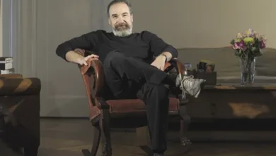 Mandy Patinkin seated in a vintage chair, relaxed and thoughtful, in a warm portrait setting