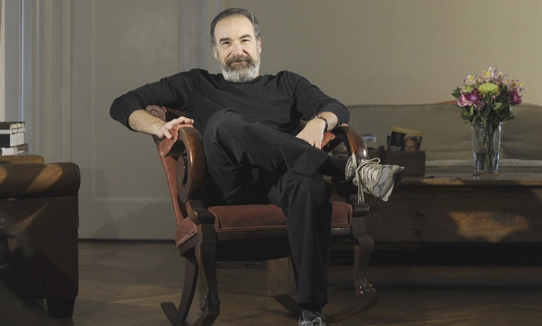 Mandy Patinkin seated in a vintage chair, relaxed and thoughtful, in a warm portrait setting