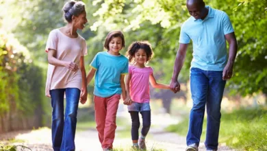 A grandmother and grandfather walking outdoors holding hands with their two grandchildren on a tree-lined path on a sunny day.