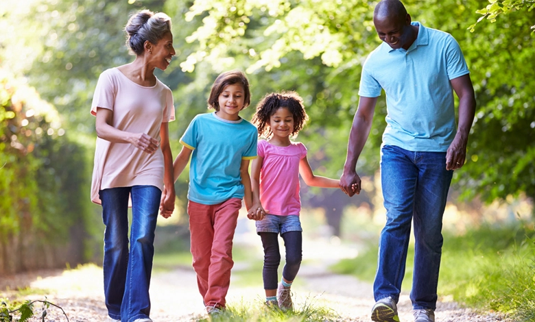 A grandmother and grandfather walking outdoors holding hands with their two grandchildren on a tree-lined path on a sunny day.