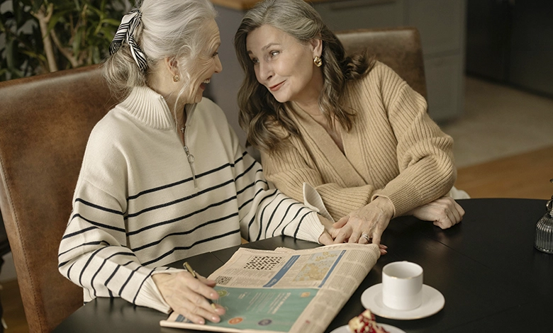 Two older women laughing together over a crossword puzzle and coffee at a café table