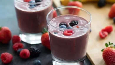 Two glasses of berry breakfast smoothie topped with fresh blackberries, blueberries, and raspberries, surrounded by strawberries and a banana.