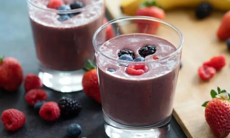 Two glasses of berry breakfast smoothie topped with fresh blackberries, blueberries, and raspberries, surrounded by strawberries and a banana.