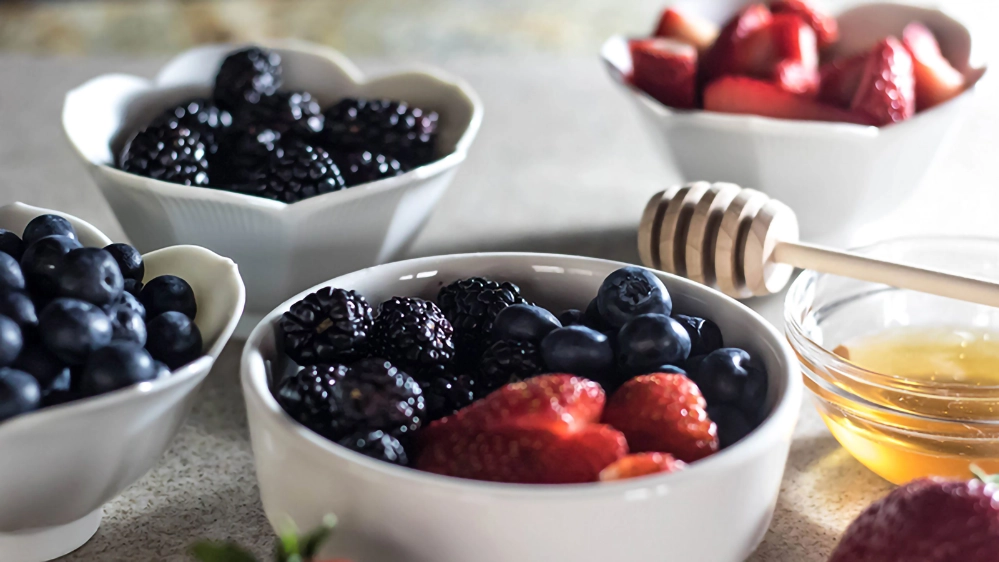 A Berry Good Breakfast Smoothie — Food for Thought 2 White bowls of fresh strawberries, blackberries, and blueberries beside a honey dipper.