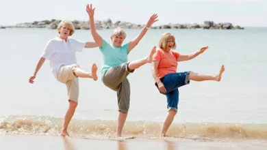 Three women laughing and dancing barefoot on the beach with arms raised in joyful celebration