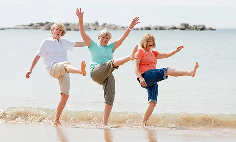 Three women laughing and dancing barefoot on the beach with arms raised in joyful celebration