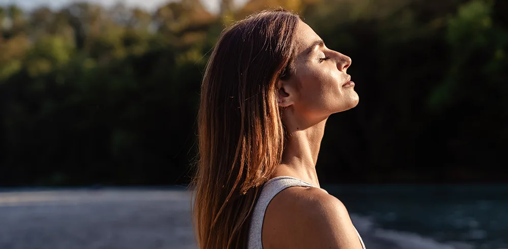 How to Boost Your Vitamin D Levels Naturally—All Year Long 3 Woman with long hair sitting in the sun, head tilted upward, and eyes closed, enjoying natural sunlight.