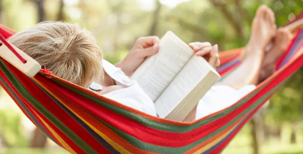 A woman around age 60 reading in a colorful outdoor hammock, her face unseen, enjoying a quiet moment in nature.
