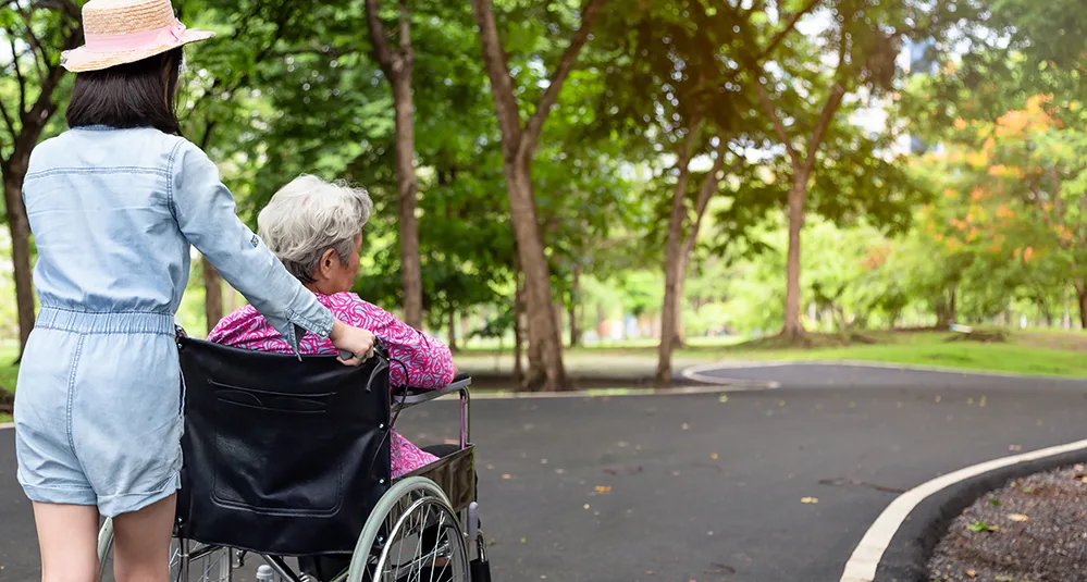 Grandparents Raising Grandchildren 2 Granddaughter pushing a senior woman in a wheelchair along a park path, symbolizing family caregiving.