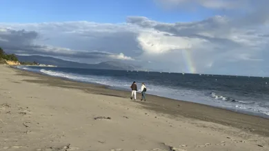 Permission to Be Unproductive 2 Two people walking side by side along the beach beneath a soft rainbow over the ocean, with waves lapping the shore and boats visible in the distance.