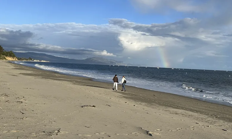 Permission to Be Unproductive 1 Two people walking side by side along the beach beneath a soft rainbow over the ocean, with waves lapping the shore and boats visible in the distance.
