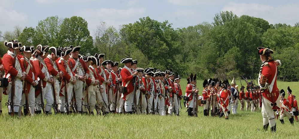 Explore Brandywine Valley: Gardens, Art, Covered Bridges & History 2 Revolutionary War reenactors in red coats lined up in formation at Brandywine Battlefield on a sunny day with green grass and trees.