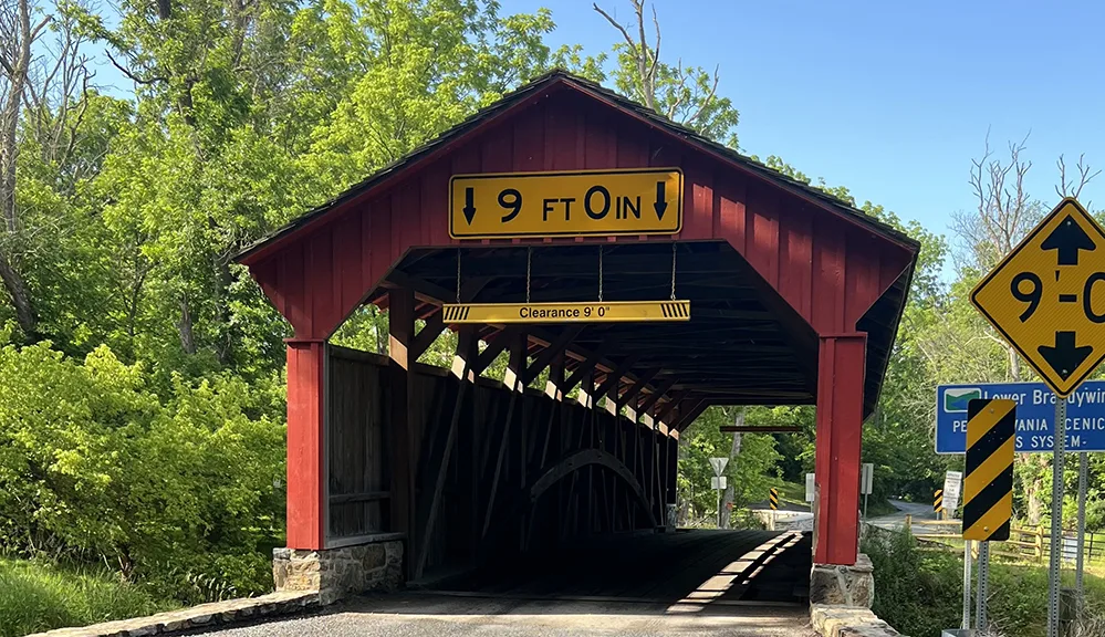 Explore Brandywine Valley: Gardens, Art, Covered Bridges & History 5 Frank Speakman Covered Bridge in Brandywine Valley, Pennsylvania, with red wood siding, historic charm, and scenic forest surroundings on a sunny day.