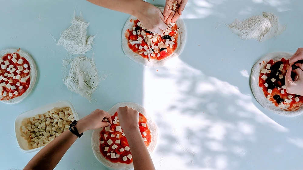 Several pairs of hands topping small pizzas with cheese and vegetables at a floured table.