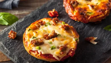 Mini pizza bagels topped with melted cheese, sausage, and green peppers on a slate board.
