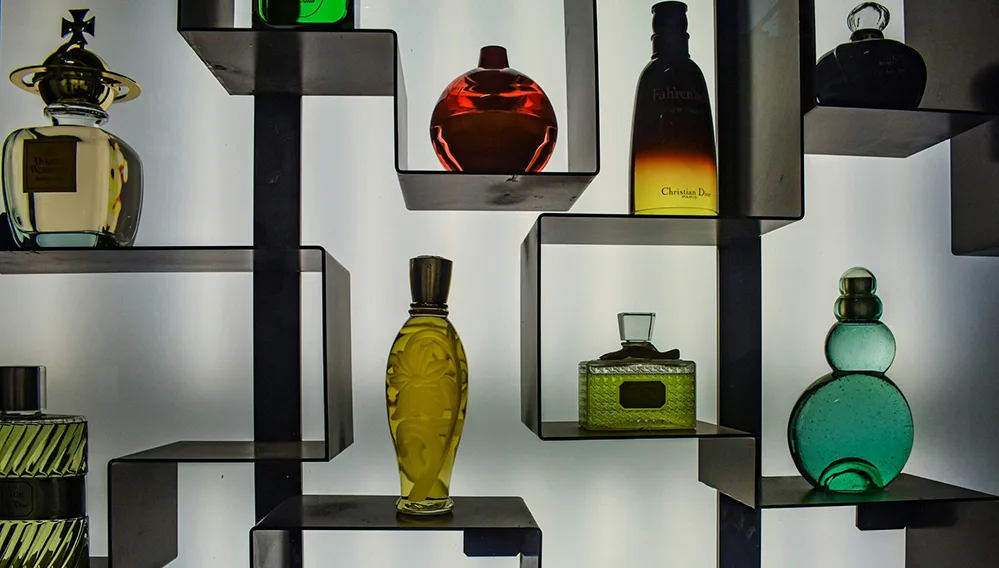 Top 10 Summer Colognes for Men 2025 2 Luxury cologne bottles in various shapes and colors displayed on black geometric shelves, featuring designer fragrances including Christian Dior and other premium brands in amber, green, and clear glass bottles