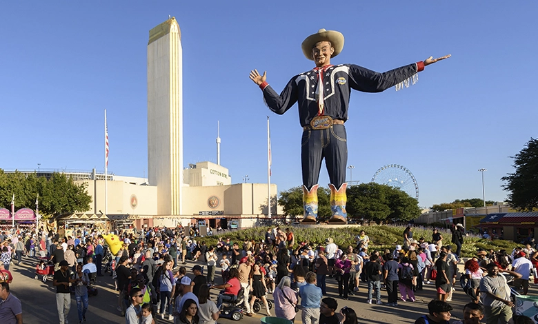 State Fair of Texas 2025 1 Aerial view of Big Tex statue surrounded by large crowds at State Fair of Texas with Cotton Bowl and Hall of State buildings in the background during golden hour.