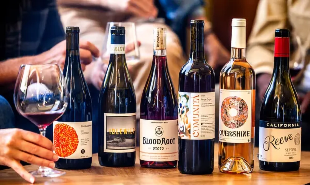 Wine Lovers 3 Six wine bottles from Overshine Collective member wineries displayed on wooden table with wine glasses and hands reaching for tasting, showing BloodRoot, Comunità, Overshine, and Reeve wine labels