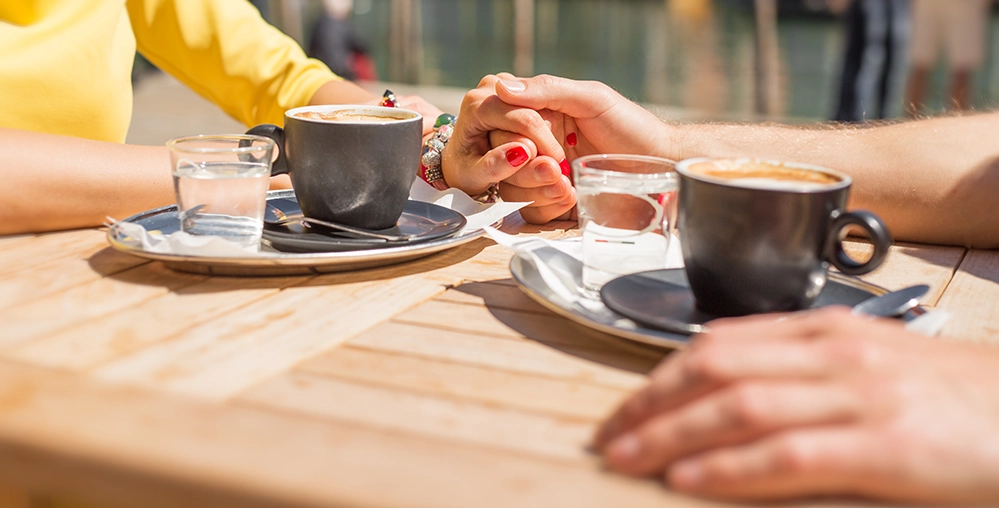 6 Secrets to Long-Term Relationships That Last After 50 3 Couple holding hands over coffee at a table, showing intimate quality time in a long-term relationship.