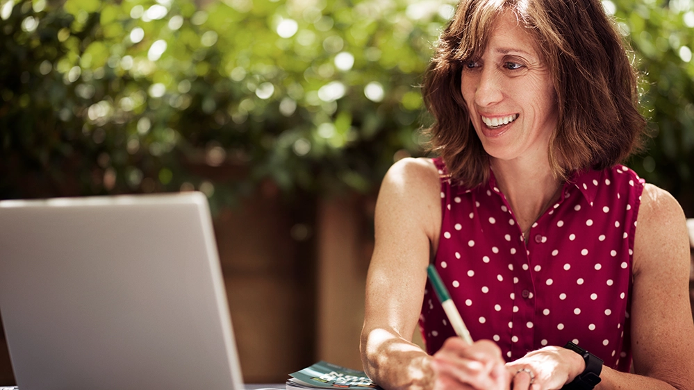 How Facebook Groups Are Helping Parents Navigate the Empty Nest Transition 2 Smiling woman in red polka dot sleeveless top taking notes on a parent group chat about college transition.