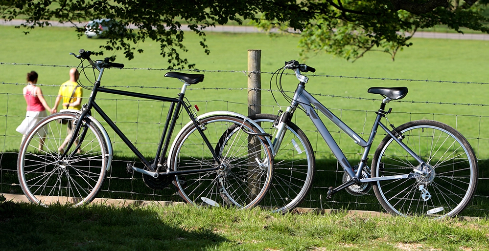 6 Secrets to Long-Term Relationships That Last After 50 4 Two bicycles parked side by side against a wooden fence with a couple walking together in the background, illustrating shared activities that keep relationships strong after 50.