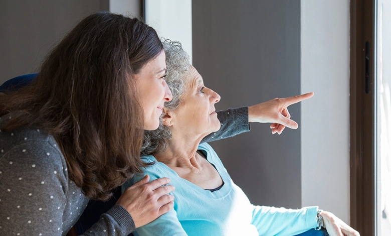 Adult daughter standing close behind elderly mother in wheelchair pointing forward together showing dementia caregiving support and connection