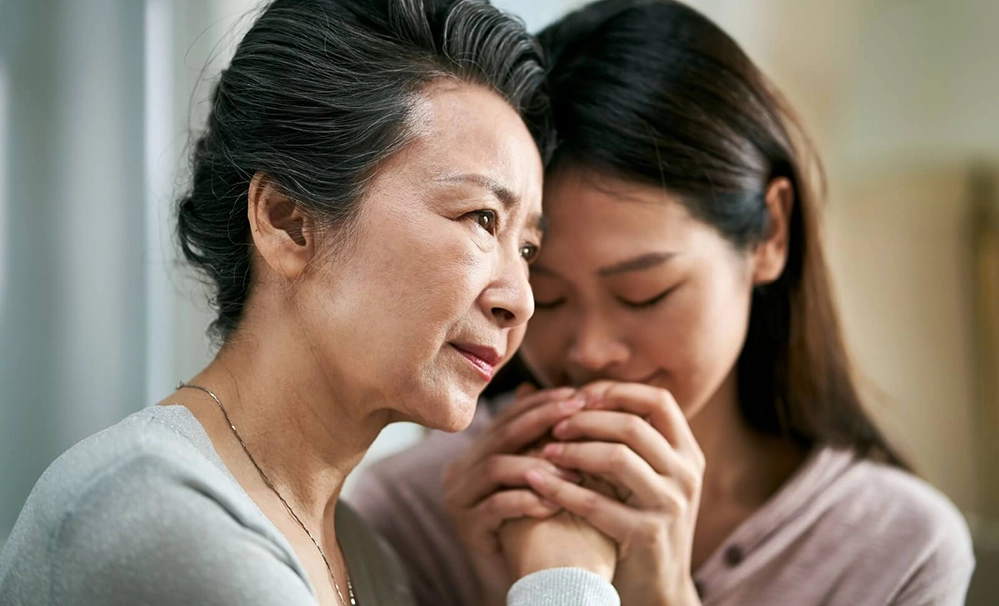 Asian daughter gently holding elderly mother's hands showing emotional support and comfort in dementia caregiving