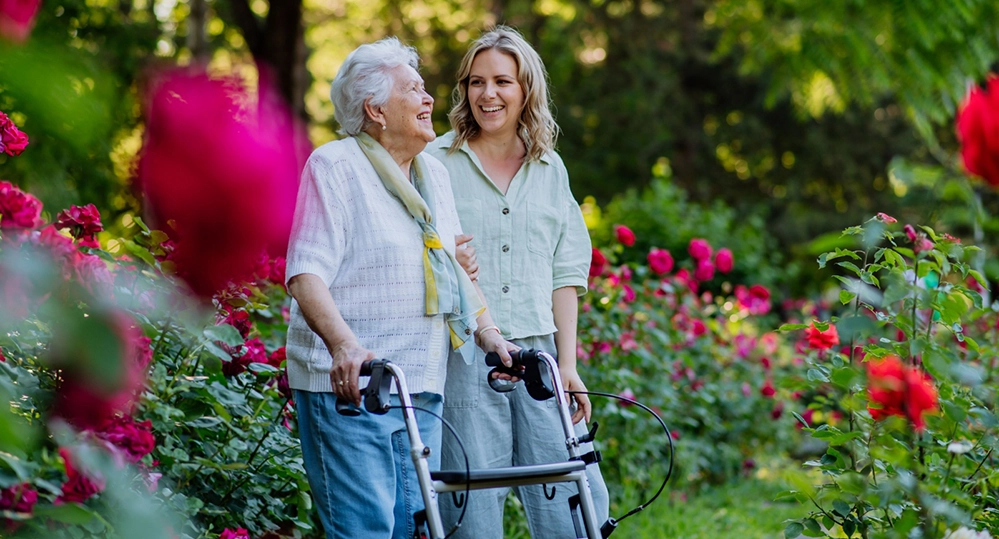 Elderly woman using walker with adult daughter caregiver walking together through colorful garden showing joy and mobility in dementia care