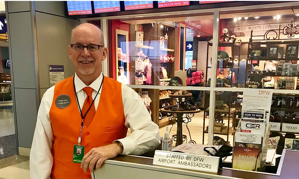 DFW Airport Ambassador standing at the information podium with flight display and shop in the background.