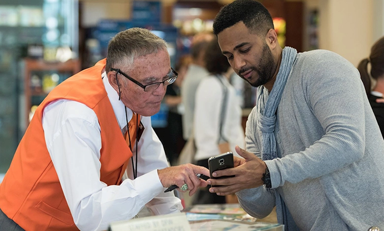 DFW Airport Ambassador in an orange vest helps a male traveler with a mobile device at the airport information center.