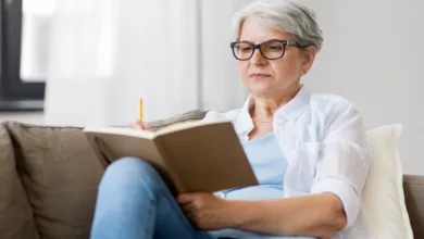 Mature woman with white hair and glasses reading a journal while relaxing on couch, practicing mindfulness for mental health in retirement
