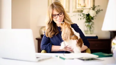 A woman in her fifties wearing glasses smiles at her laptop while holding a Cavalier King Charles Spaniel in her arms at a home desk, a reminder that pets support brain health after 50.