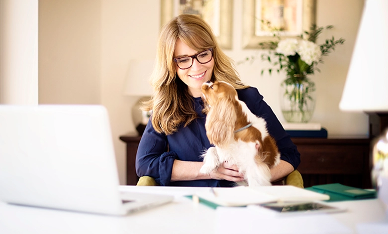 A woman in her fifties wearing glasses smiles at her laptop while holding a Cavalier King Charles Spaniel in her arms at a home desk, a reminder that pets support brain health after 50.