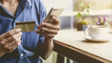 Older woman in blue shirt holding credit card and smartphone making online purchase at home