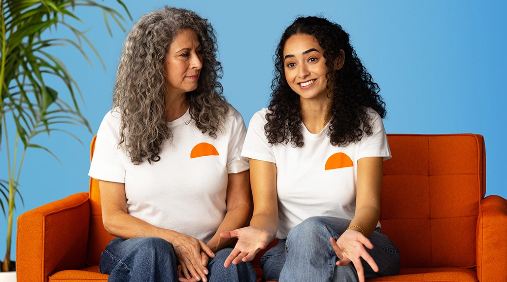 Two women from different generations wearing Uqora branded shirts sitting together on orange couch representing intergenerational approach to women's urinary health