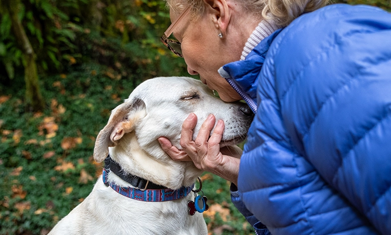 Older woman with glasses tenderly kissing her white lab mix dog outdoors, showing the deep emotional bond between pets and humans.