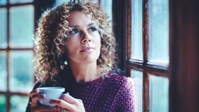 A woman holding a cup of coffee, gazing out a window in thought