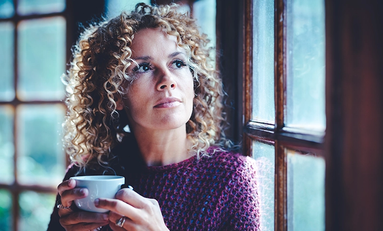 A woman holding a cup of coffee, gazing out a window in thought