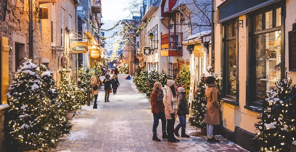 Snow-covered cobblestone street in Old Québec City with glowing lights, decorated trees, and people strolling.