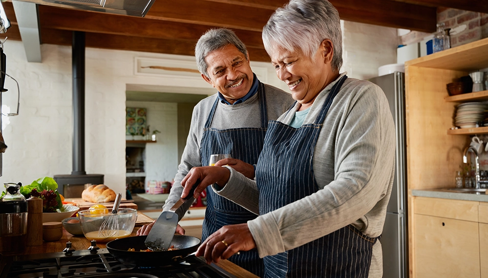 Planning to Retire? 2 Smiling multiethnic senior couple cooking breakfast together in their kitchen, symbolizing joy, connection, and new possibilities in retirement.