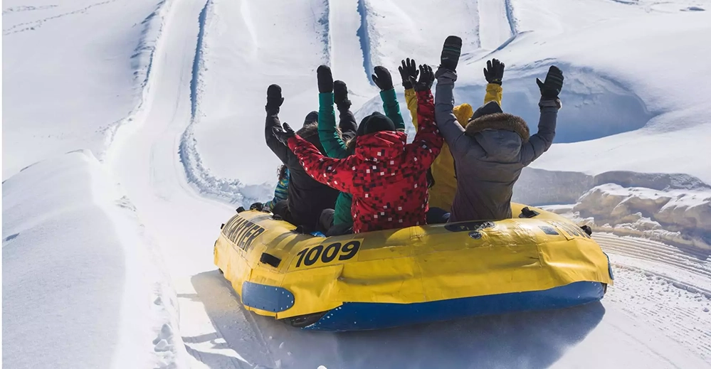 Group of bundled-up riders raising their arms while snow rafting at Village Vacances Valcartier near Québec City, surrounded by snowy hills.