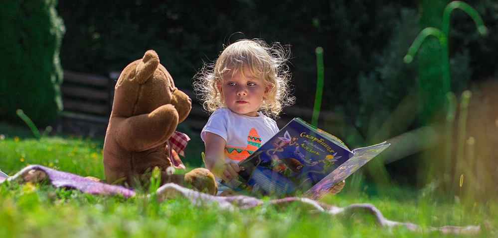 Young toddler girl reading book outdoors with teddy bear showing independent love of reading developed from infancy