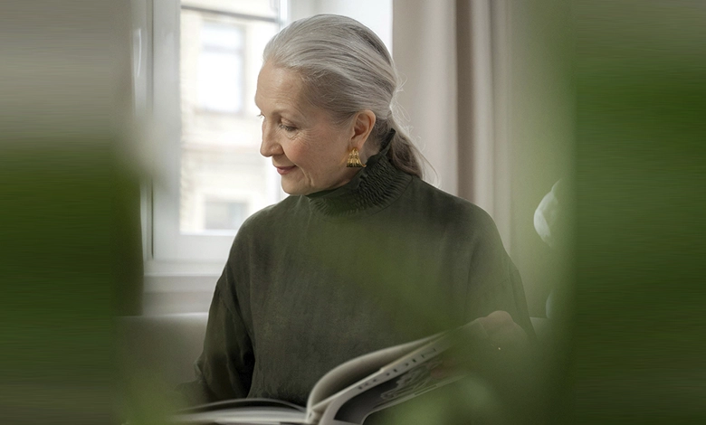 Woman with silver hair reading a book by a window as part of her self-care routine