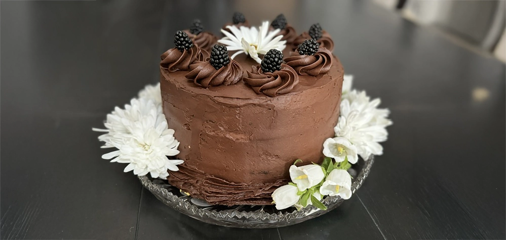 Chocolate-mayonnaise cake with smooth chocolate frosting, piped rosettes topped with blackberries, and white flowers arranged around the base on a glass cake stand