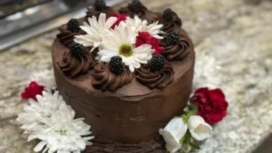 Three-layer chocolate-mayonnaise cake with chocolate frosting, piped rosettes, fresh berries, white daisies, and red carnations on a glass cake stand