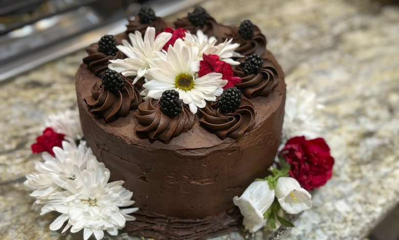 Three-layer chocolate-mayonnaise cake with chocolate frosting, piped rosettes, fresh berries, white daisies, and red carnations on a glass cake stand