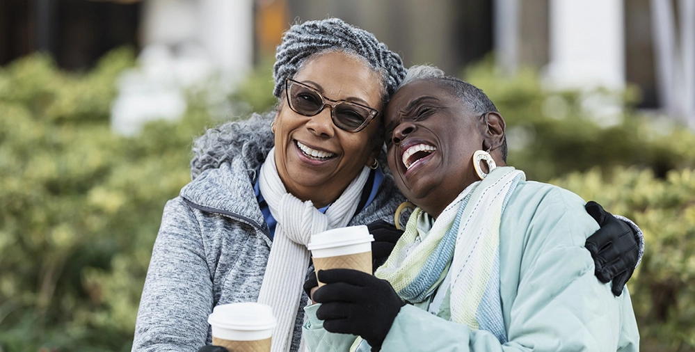 Two women in winter coats laughing together while holding coffee cups outdoors