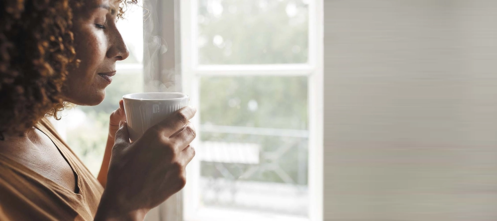 Woman in profile holding a steaming coffee cup by a sunlit window