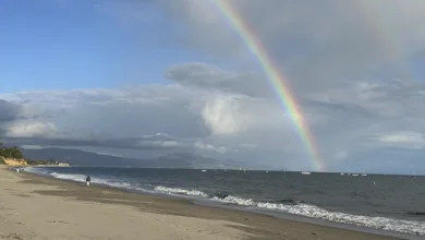 Rainbow arching over a quiet Santa Barbara beach with dramatic clouds and blue sky